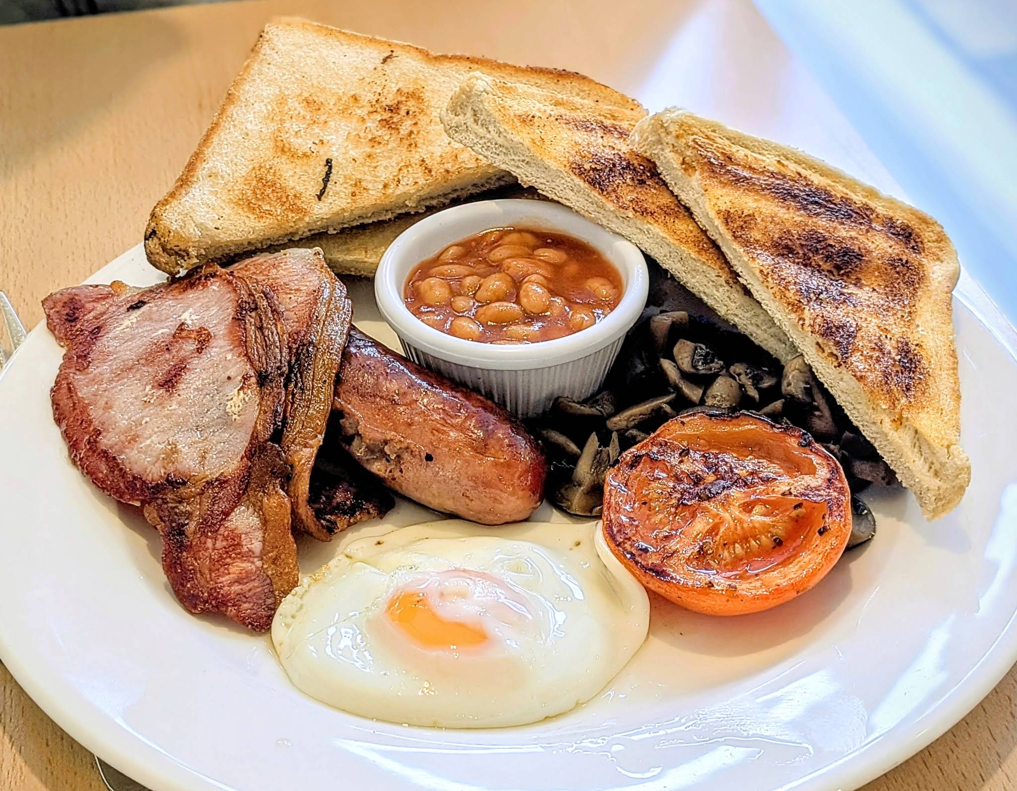 A full white breakfast plate with egg, bacon, sausage, toast, beans and tomato